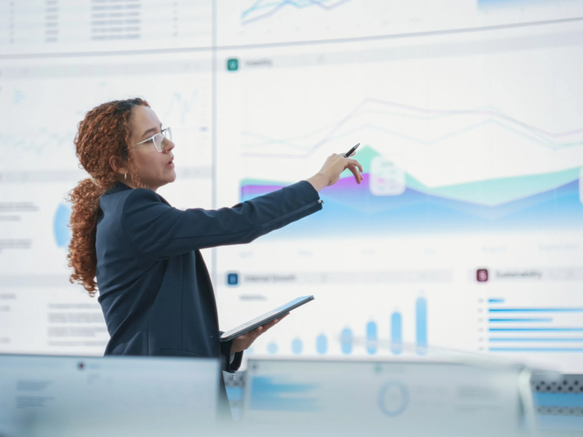 A business professional presenting data on a large digital screen, pointing to charts and graphs during a strategy or performance briefing.